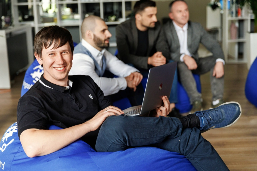 A man smiles while working on a laptop in an office setting. Three other men sit in the background.
