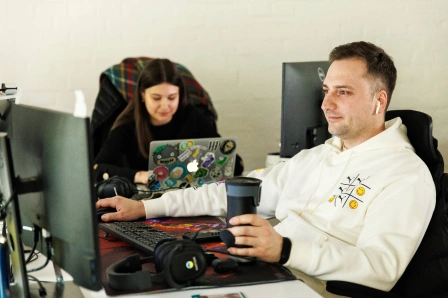 A person in a white hoodie sits at a desk with a computer, wearing earbuds and holding a travel mug. In the background, another person is focused on a laptop with colorful stickers. Headphones are on the desk.