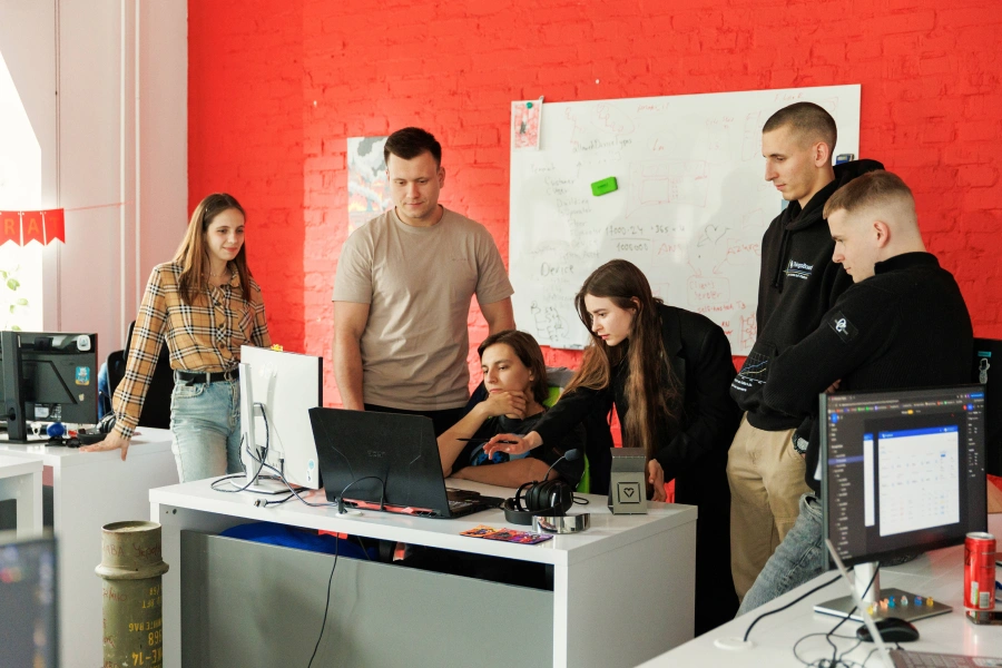 A group of six people gathered around a desk with computers and a laptop, collaborating on a project. The room has a vibrant red brick wall with brainstorming notes on a whiteboard. Everyone is focused and engaged in discussion.